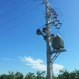 Słup energetyczny z transformatorem i osprzętem, widok z dołu na tle błękitnego nieba z lekkimi obłokami, zieleń drzew w dolnej części kadru.
