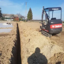 Wąski wykop w piaszczystej ziemi, obok czarna mini koparka Bobcat, w tle dom z ogrodem i błękitne niebo.
