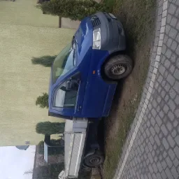Niebieski bus z otwartymi drzwiami, zaparkowany na kostce brukowej, widok na budynek w tle.
