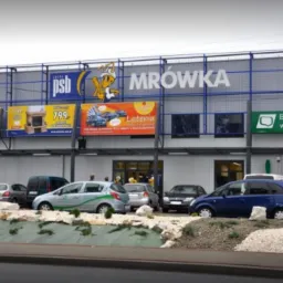 Fasada sklepu budowlanego Mrówka z reklamami na górze, parking przed sklepem z samochodami i kamieniami ozdobnymi.