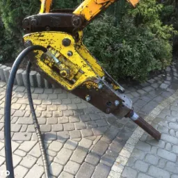 Żółty młot hydrauliczny z widocznymi śladami użytkowania i przewodami hydraulicznymi, ustawiony na kostce brukowej w pobliżu zieleni.