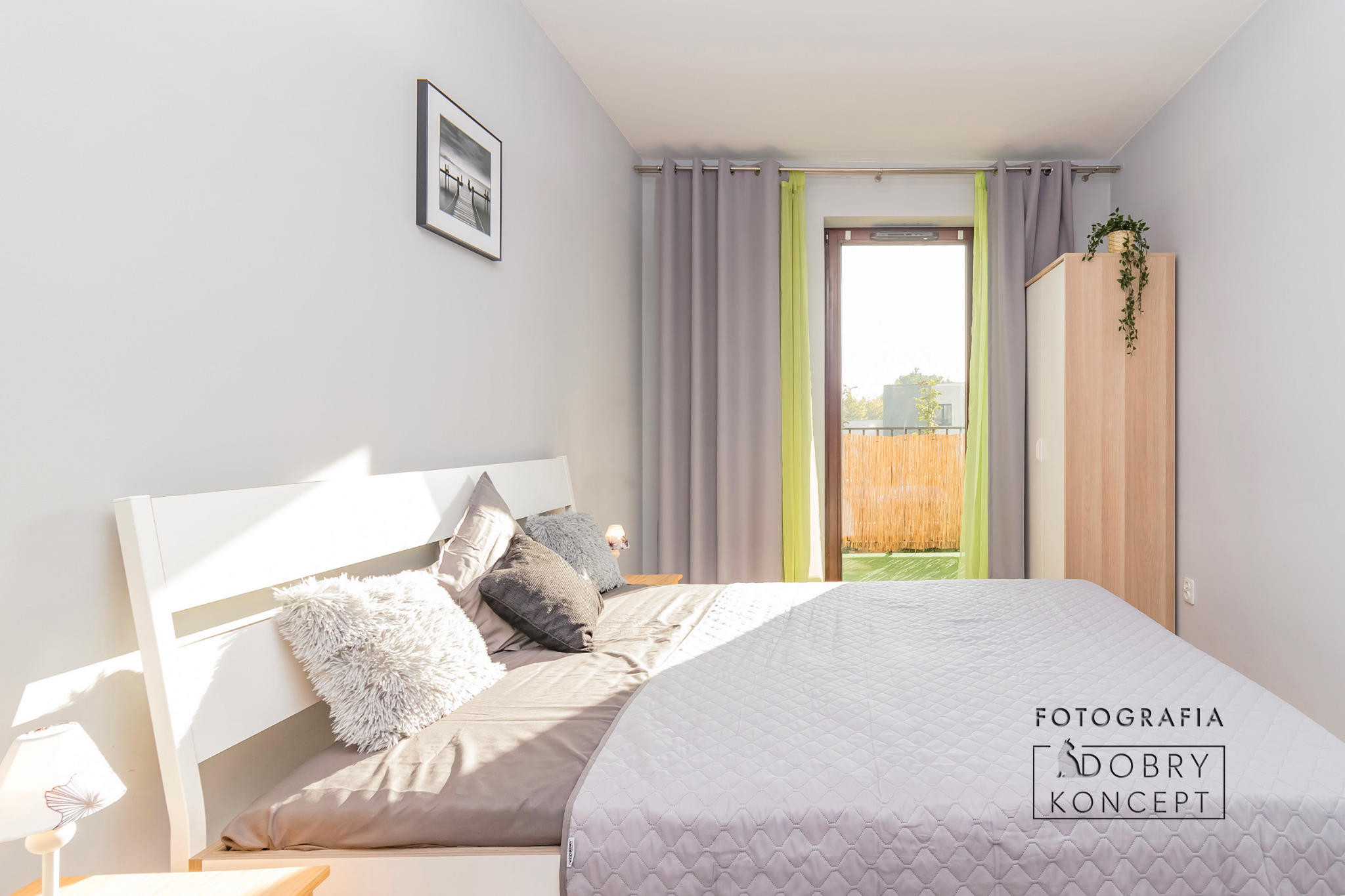 Jasna sypialnia z białym łóżkiem, puszystymi poduszkami, szarymi zasłonami i zielonym akcentem w postaci firanki, z widokiem na balkon z bambusową osłoną.