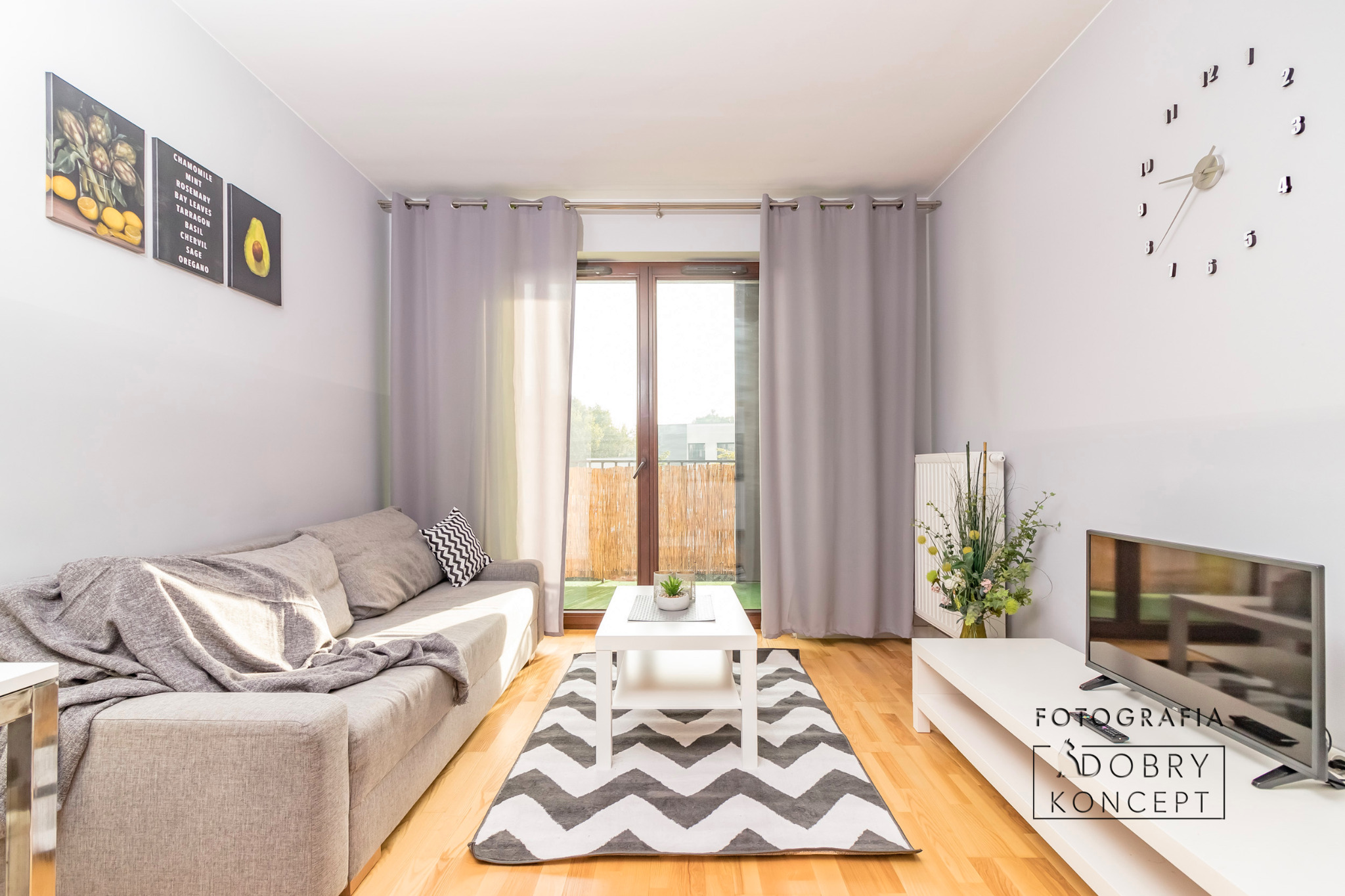 Jasne wnętrze salonu z szarą sofą, białym stolikiem kawowym na dywanie w zygzaki, telewizorem i dekoracjami ściennymi.