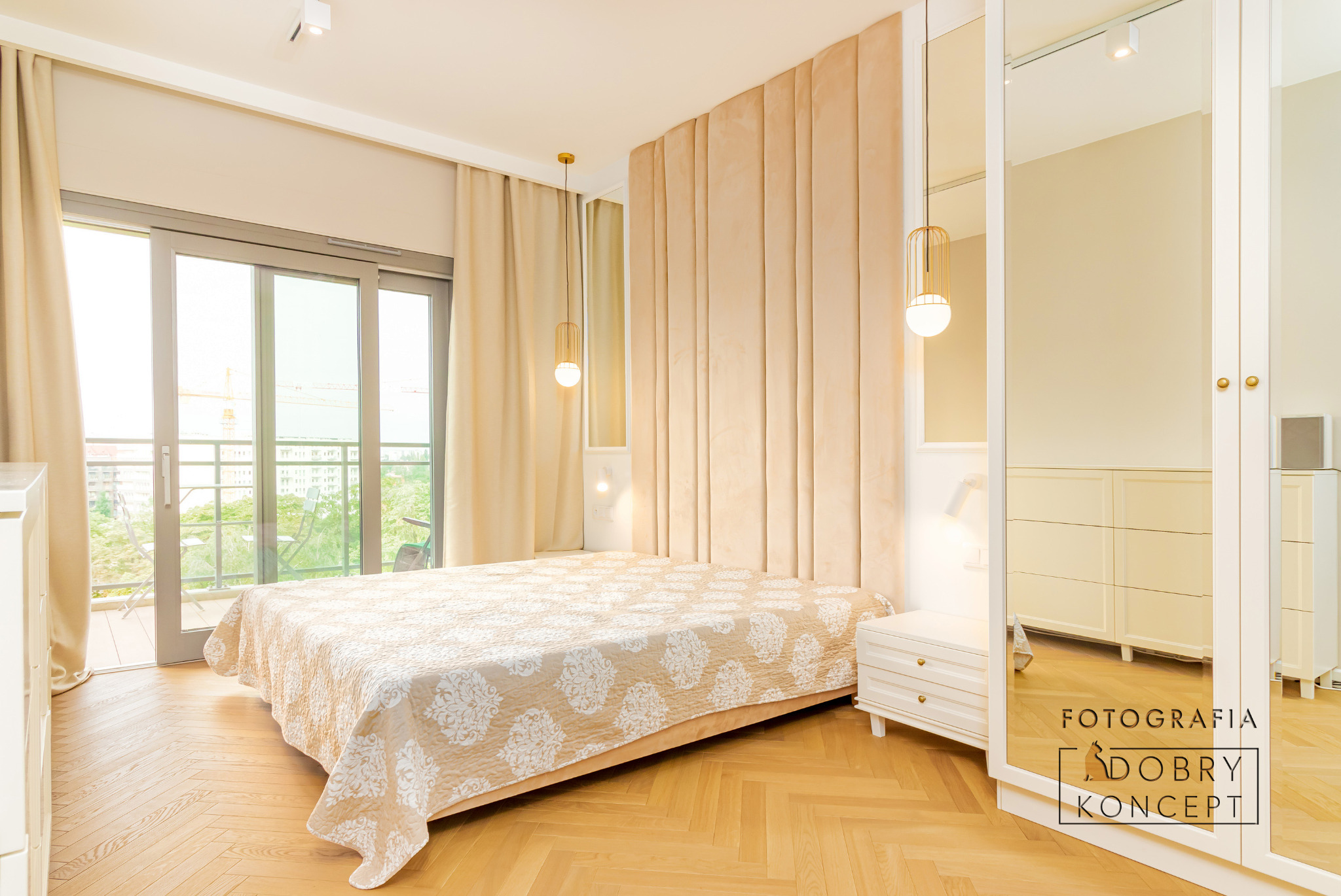 Jasna sypialnia z łóżkiem, beżowym tapicerowanym zagłówkiem, parkietem w jodełkę i wyjściem na balkon z widokiem na zieleń miejską. W rogu lustrzana szafa z widocznym logo firmy.