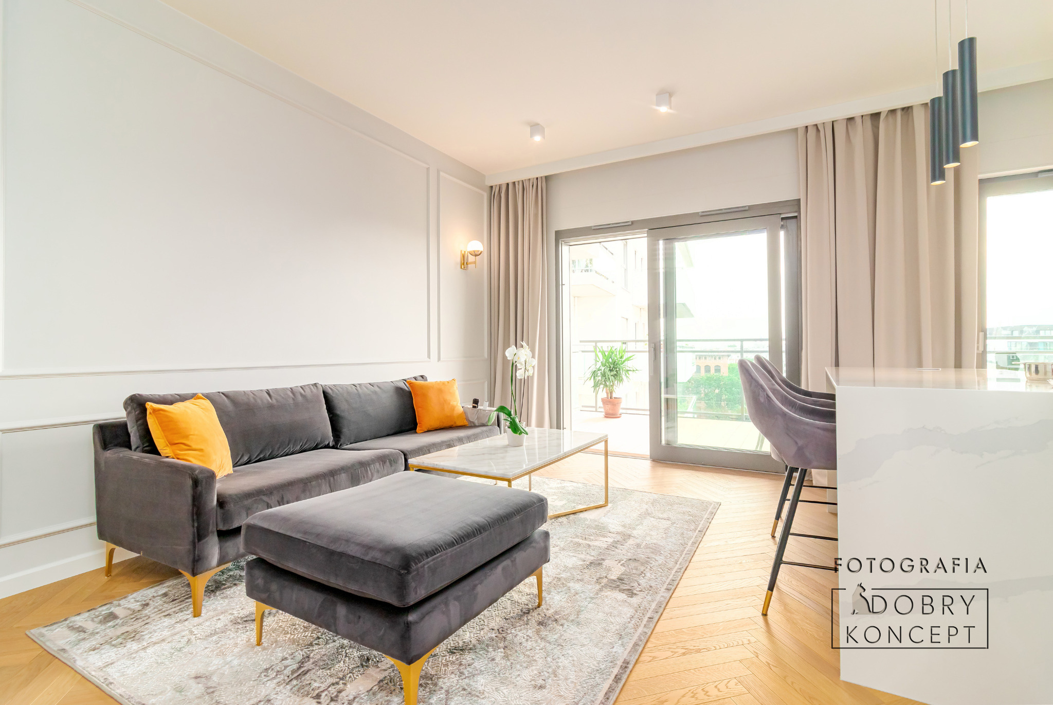 Jasne wnętrze salonu we Wrocławiu z szarą sofą narożną, pufem, marmurowym stolikiem kawowym, dywanem w odcieniach szarości i złotymi akcentami, balkon z widokiem na miasto.