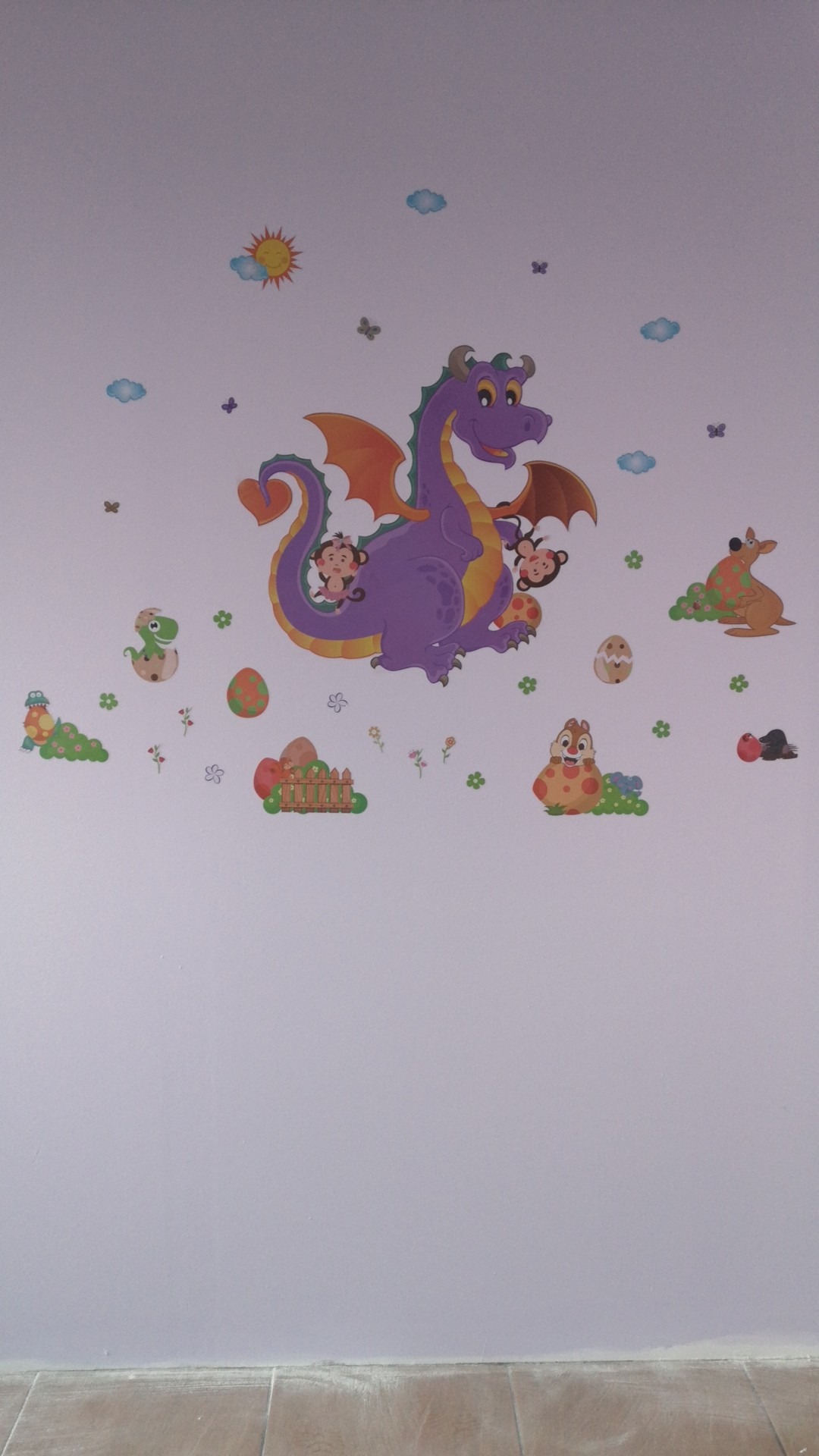 Ściana pokoju dziecięcego ozdobiona naklejkami ze smokiem, małpkami, słońcem, chmurkami i innymi zwierzętami, tworząc radosny i kolorowy motyw.