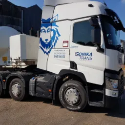 Biała ciężarówka z naczepą-cysterną firmy Gonika Trans, z logo wilka, zaparkowana na placu, widoczne zbiorniki w tle.