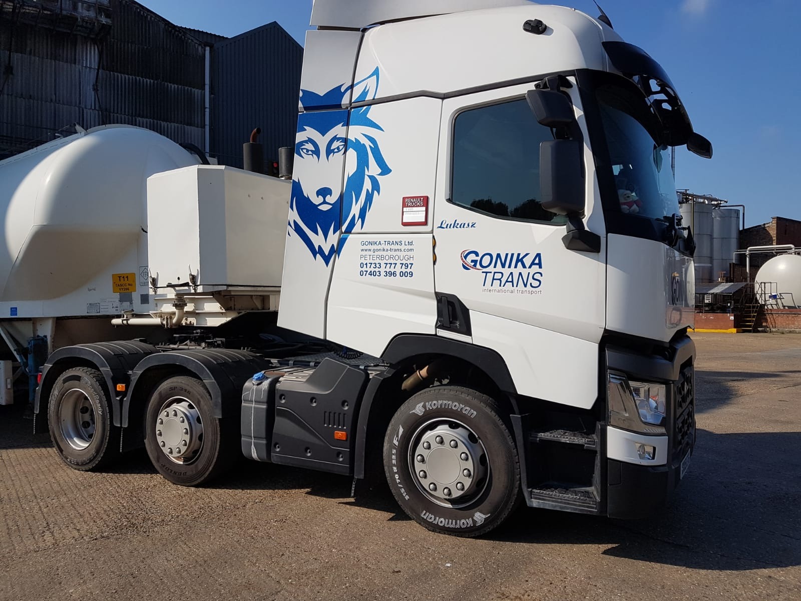 Biała ciężarówka z naczepą-cysterną firmy Gonika Trans, z logo wilka, zaparkowana na placu, widoczne zbiorniki w tle.