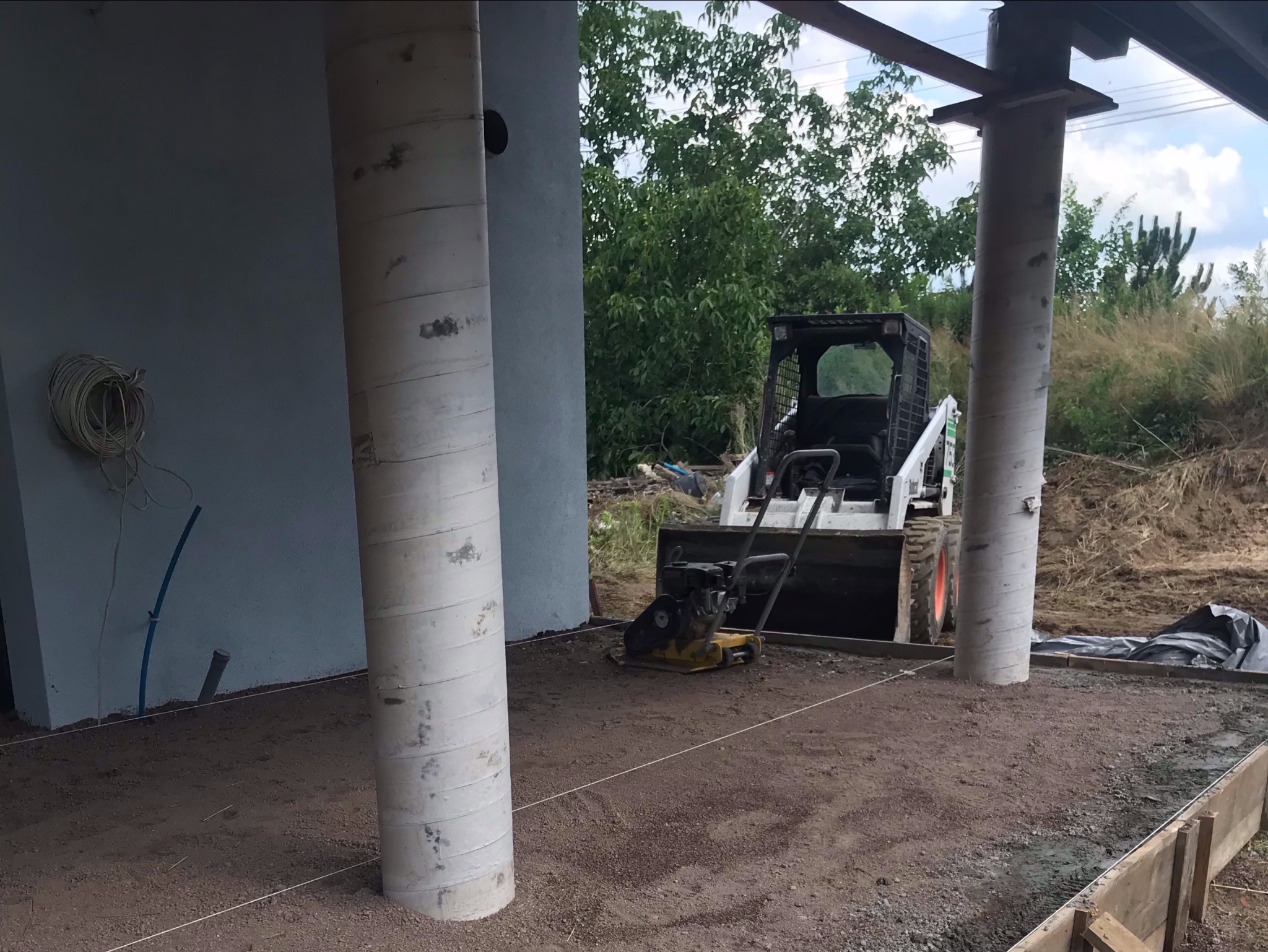 Przygotowanie podłoża pod nawierzchnię: ubijarka wibracyjna i miniładowarka na gruncie stabilizowanym, widoczne sznurki wyznaczające poziom, kolumny konstrukcyjne budynku w tle.