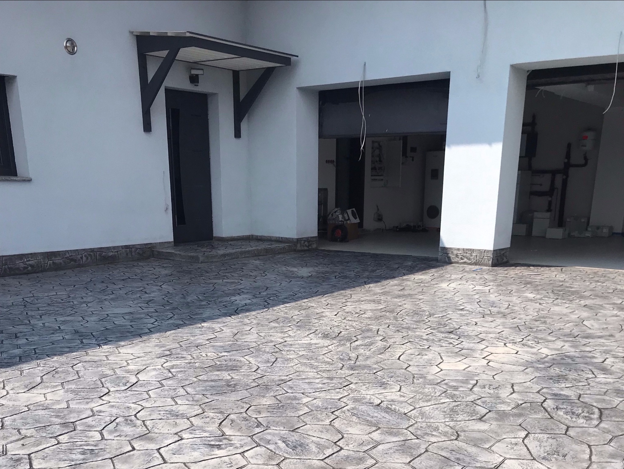 Wykończony podjazd z szarej kostki imitującej kamień, widok na wejście do garażu i drzwi wejściowe z zadaszeniem.