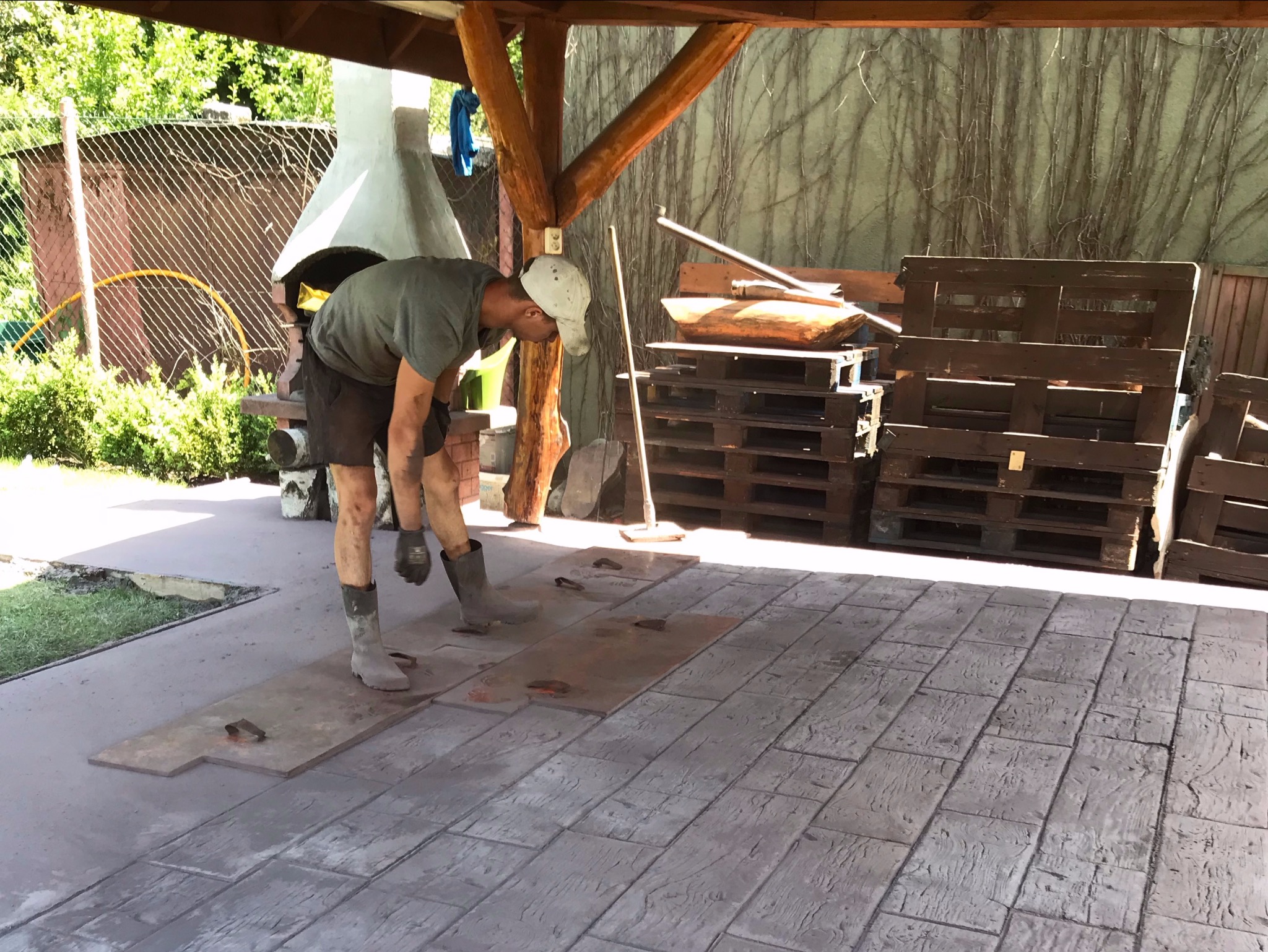 Pracownik w gumowych butach i roboczych rękawicach pochyla się nad formami do odciskania wzoru desek na świeżo wylanej betonowej posadzce tarasu, w tle palenisko i palety.