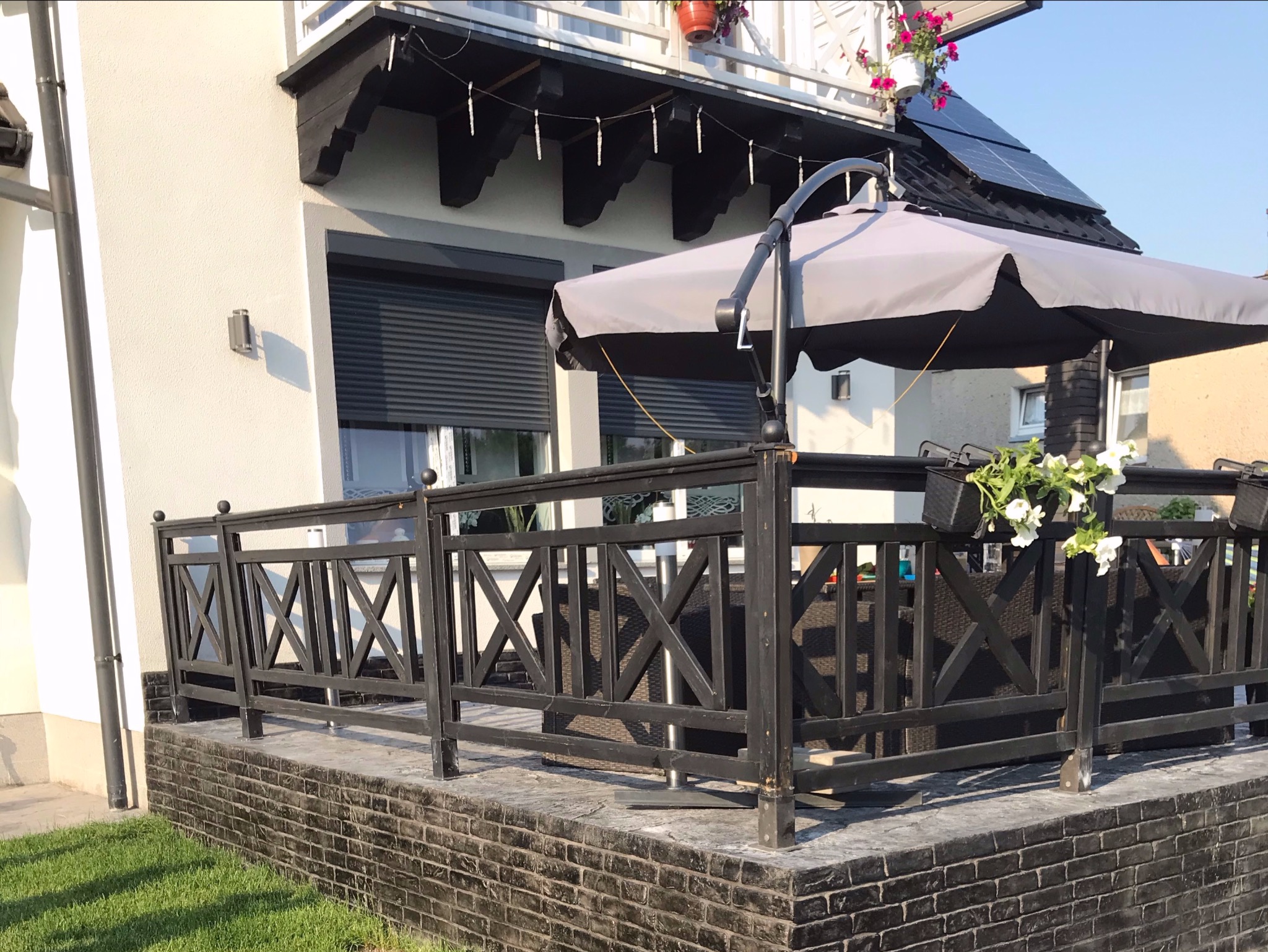 Zadaszony taras z czarną, drewnianą balustradą i szarym parasolem, wykończony kostką imitującą cegłę, z widocznymi donicami z kwiatami.