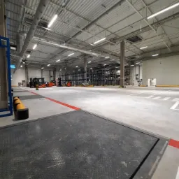 Wnętrze hali magazynowej z widocznymi lampami przemysłowymi, regałami i wózkami widłowymi w oddali, oświetlone jasnym, sztucznym światłem.