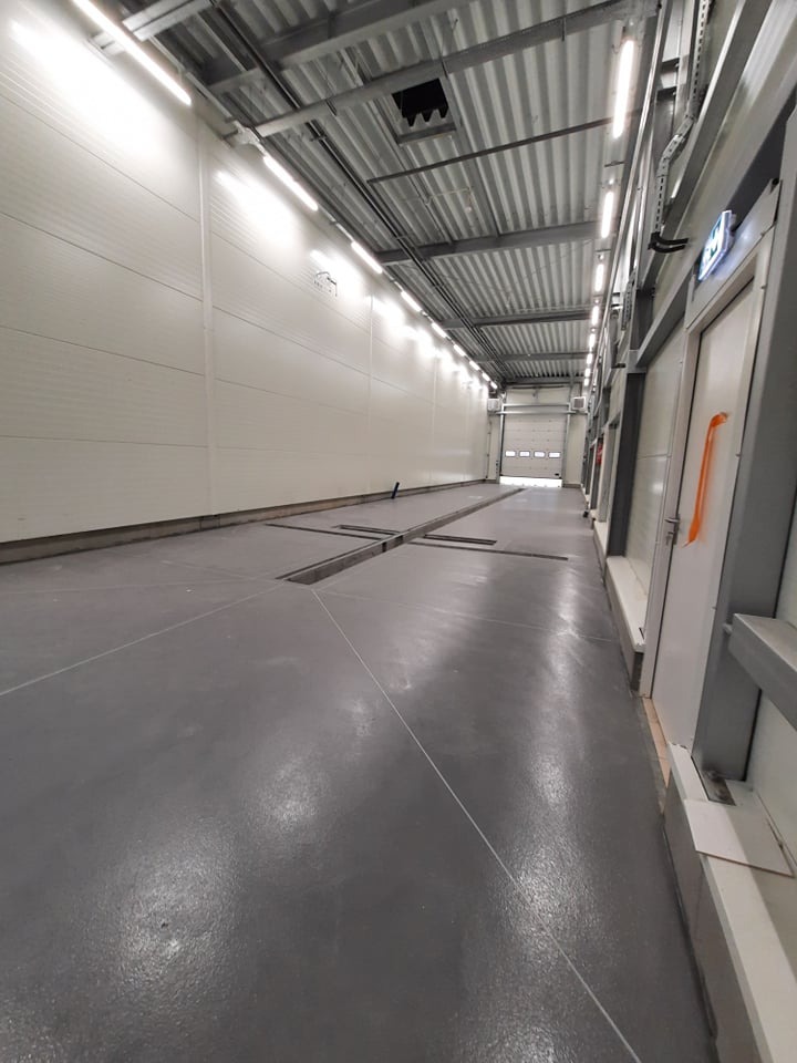 Wnętrze hali produkcyjnej z szarym betonowym podłożem, kanałami rewizyjnymi, białymi ścianami z paneli i rzędem świetlówek pod sufitem. Widoczna brama garażowa w oddali i drzwi po prawej stronie.