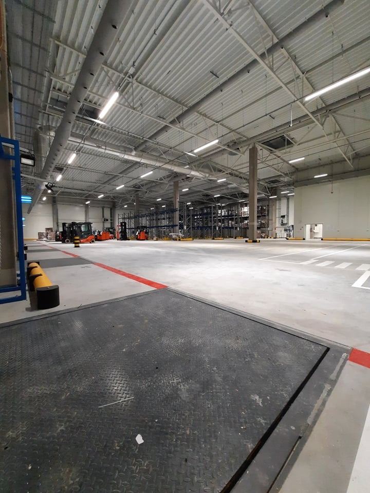 Wnętrze hali magazynowej z widocznymi lampami przemysłowymi, regałami i wózkami widłowymi w oddali, oświetlone jasnym, sztucznym światłem.