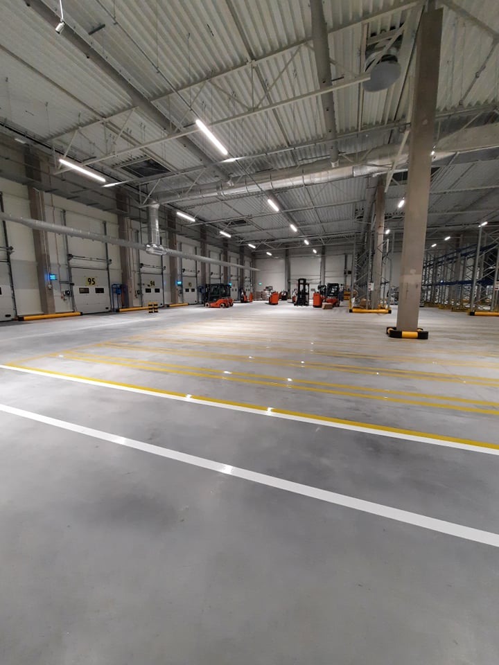 Przestronne wnętrze hali magazynowej z widocznymi liniami wyznaczającymi strefy, oświetleniem sufitowym i kilkoma wózkami widłowymi w tle.