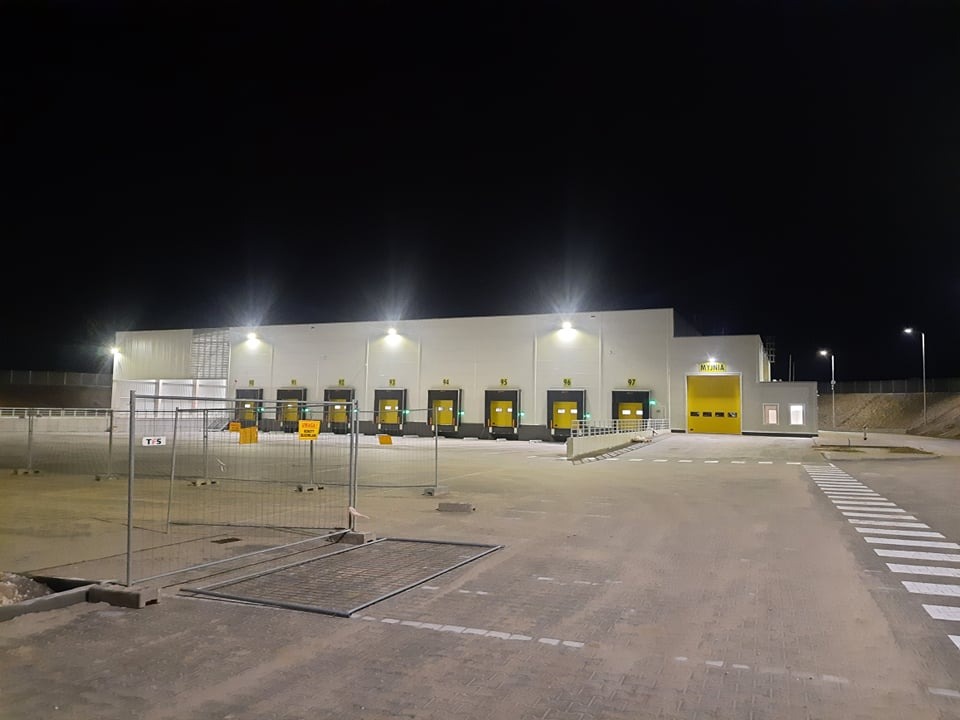 Oświetlony nocą budynek magazynowy z rampami za ogrodzeniem tymczasowym, widoczne numery ramp i lampy oświetlające elewację.