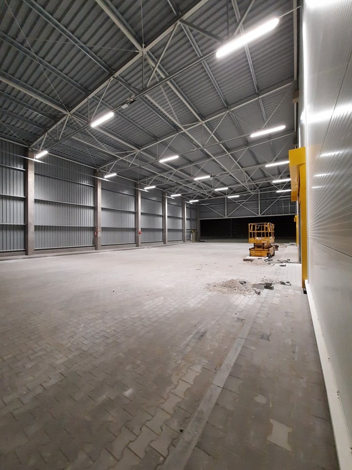 Wnętrze hali magazynowej z widoczną konstrukcją stalową dachu, oświetleniem LED, posadzką z kostki brukowej oraz żółtym podnośnikiem koszowym w tle. Fragment gruzu na posadzce.