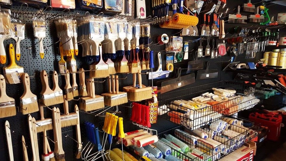 Różnorodne pędzle malarskie z drewnianymi uchwytami, wałki do malowania i szpachelki eksponowane na perforowanej ścianie w sklepie z artykułami malarskimi.