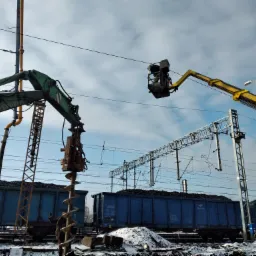 Żółty podnośnik koszowy z pracownikiem przy liniach wysokiego napięcia nad wagonami kolejowymi z węglem, obok zielony żuraw z wiertłem ślimakowym, zimowy krajobraz.