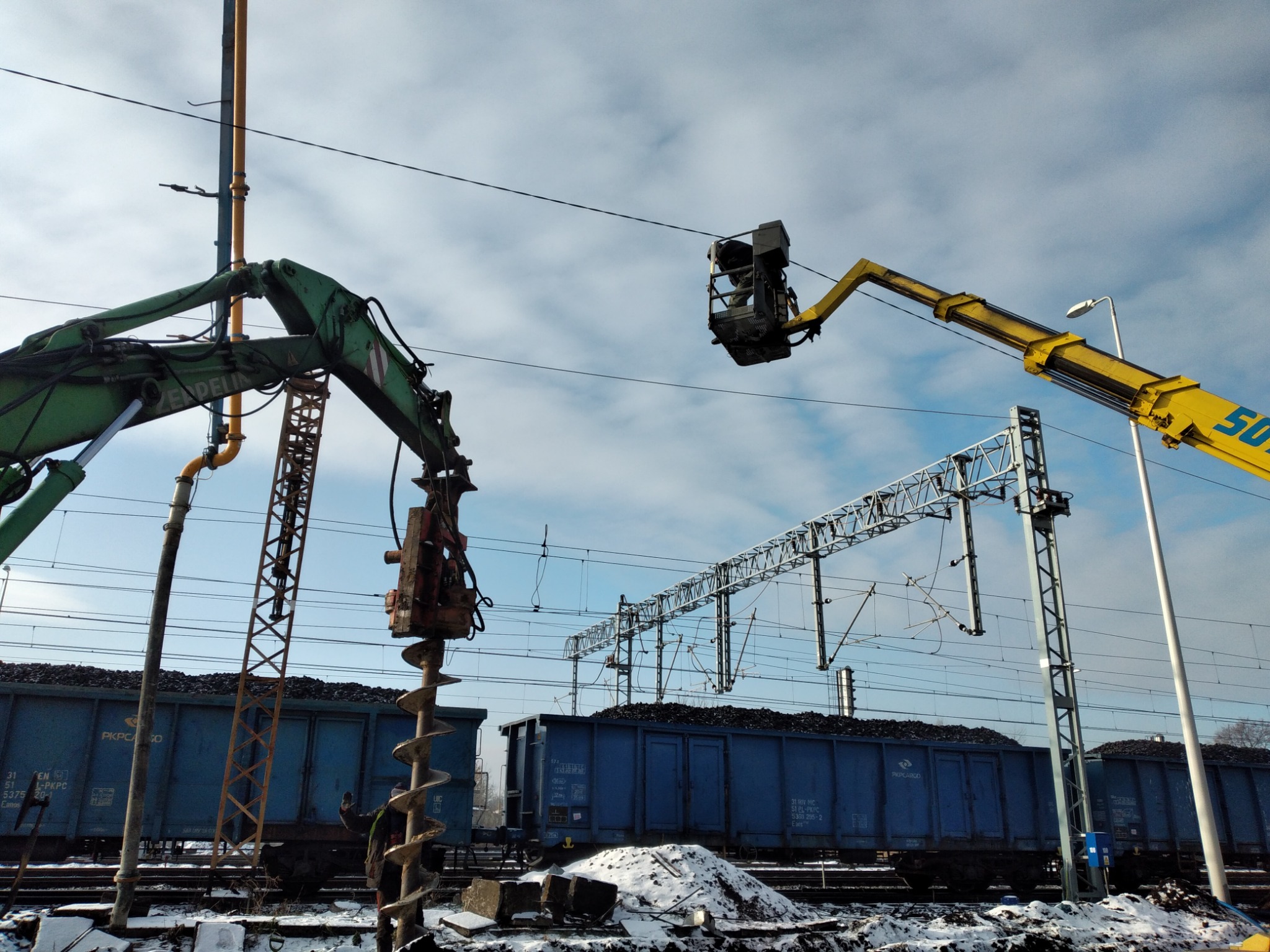 Żółty podnośnik koszowy z pracownikiem przy liniach wysokiego napięcia nad wagonami kolejowymi z węglem, obok zielony żuraw z wiertłem ślimakowym, zimowy krajobraz.