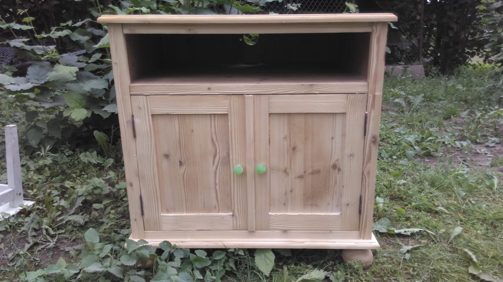 Drewniana szafka RTV z zielonymi gałkami po sodowaniu, stojąca na trawie, widoczne ślady obróbki drewna.