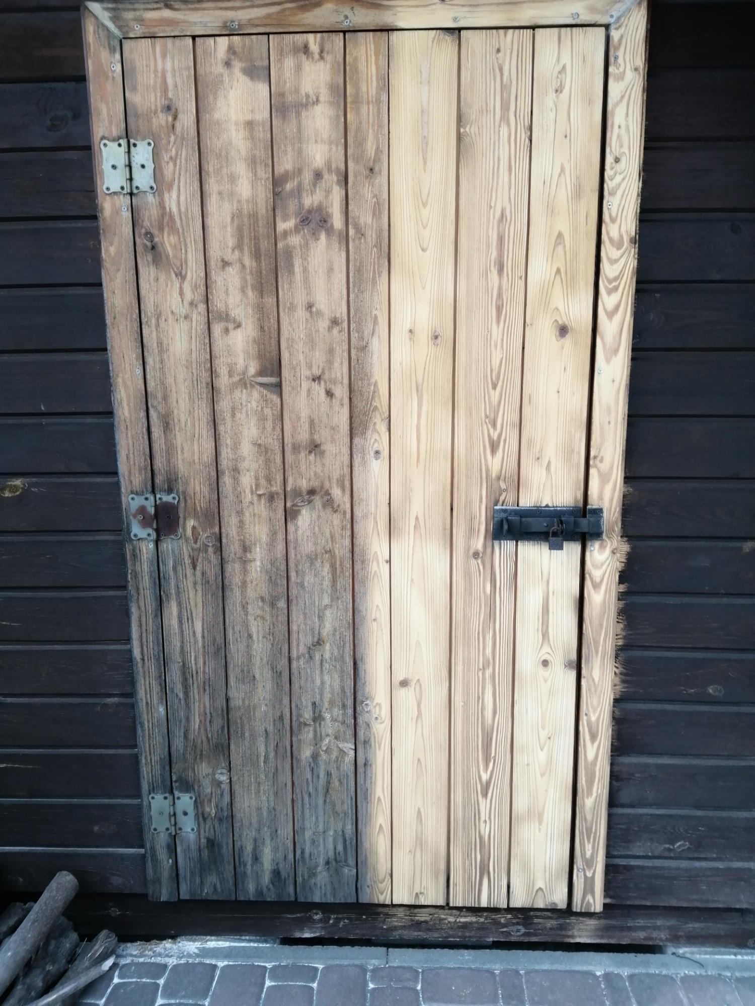 Drewniane drzwi częściowo oczyszczone, ukazujące kontrast między surowym, ciemniejszym drewnem a jasnym, odnowionym fragmentem, zawiasy z widoczną korozją, zamek na jasnej części drzwi.