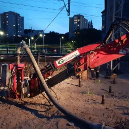 Przewiert sterowany wykonywany nocą przy torach kolejowych, widoczny czerwony sprzęt z logo Geo-Ekspert oraz robotnik w tle, w oddali oświetlone budynki mieszkalne.