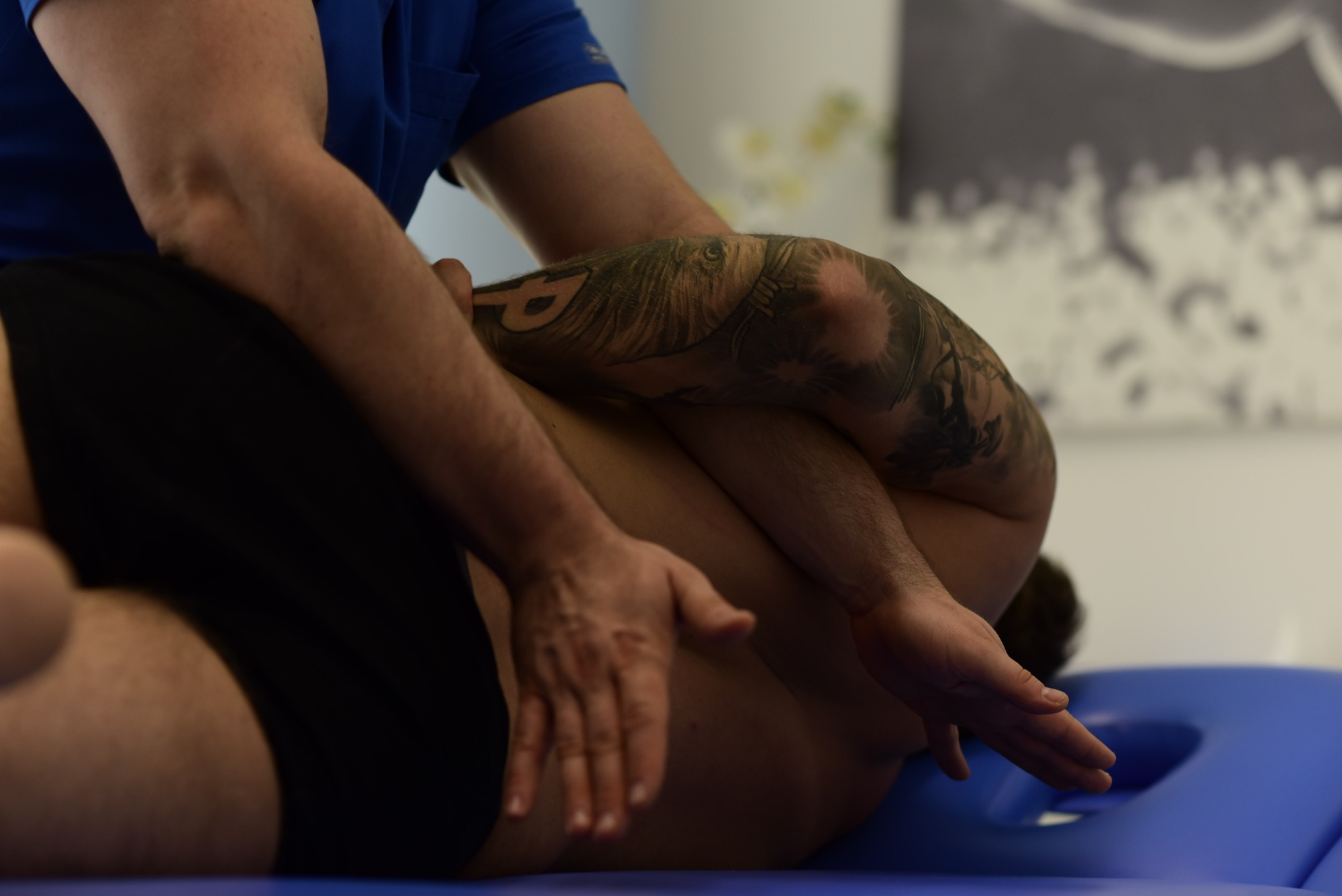 Mężczyzna w niebieskiej koszulce z tatuażem na ramieniu wykonuje masaż pleców pacjenta leżącego na niebieskim stole do masażu.