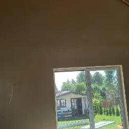 Wnętrze poddasza z nałożonymi gładziami, widoczne krokwie dachowe, przez okno widok na ogród z domkiem.