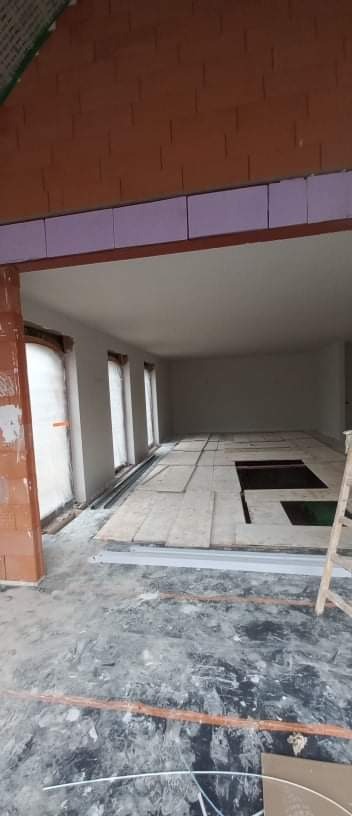 Wnętrze w trakcie prac wykończeniowych: surowy mur z cegły, izolacja, wylewka podłogowa z fragmentami płyt i drabina oparta o ścianę.