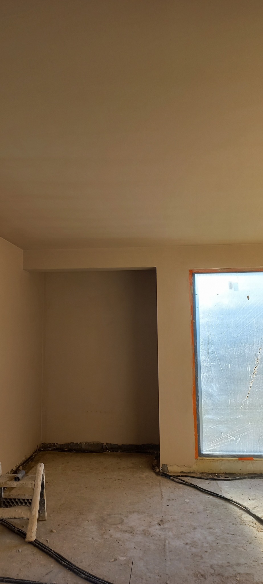 Wnętrze po nałożeniu gładzi na ścianach i suficie, widoczne okno z pomarańczowym obramowaniem i fragment drabiny malarskiej przy ścianie.