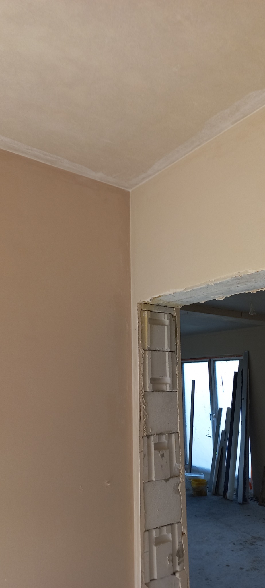 Świeżo otynkowana ściana wewnętrzna w odcieniach beżu i bieli, widoczne surowe wykończenie otworu drzwiowego z bloków betonowych, w tle okno i materiały budowlane.