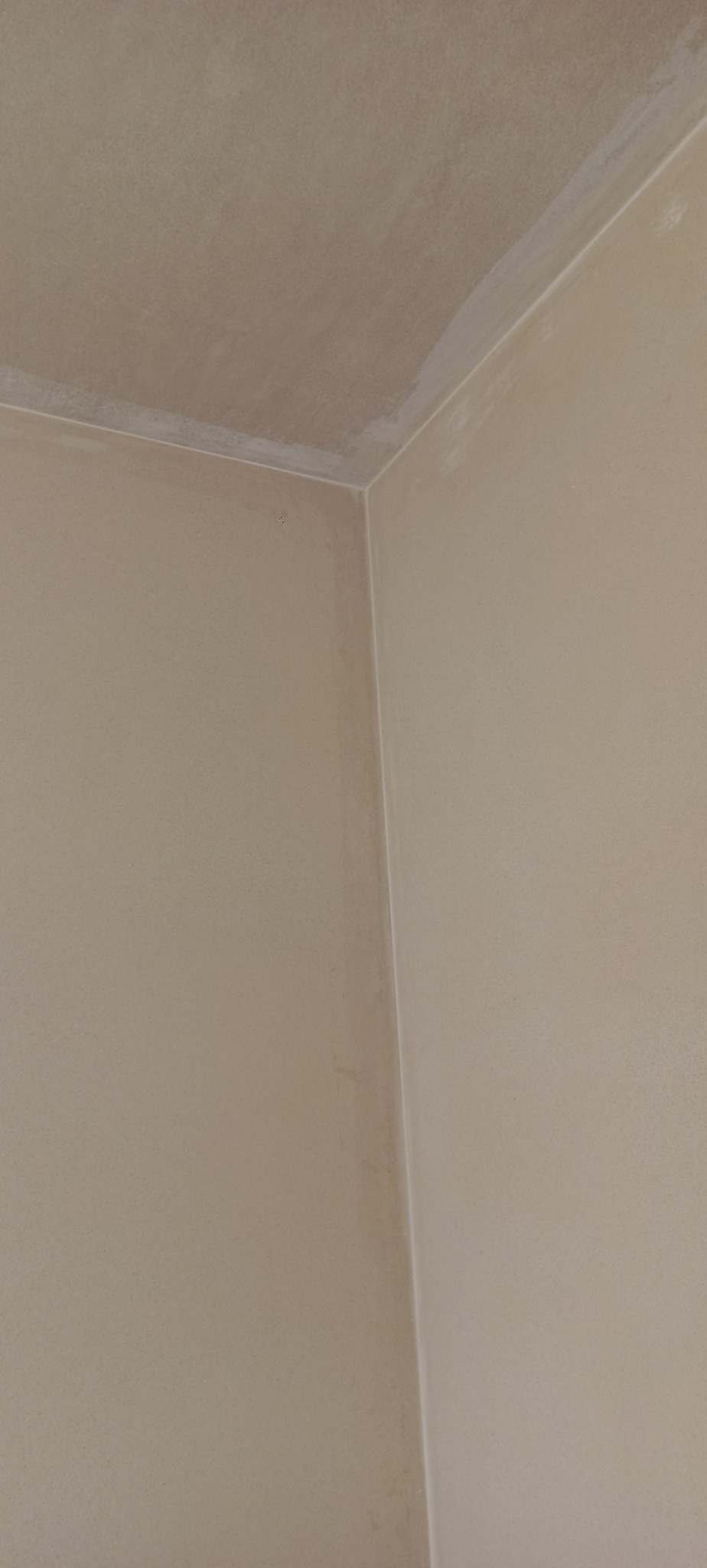 Świeżo otynkowany narożnik wewnętrzny pomieszczenia, widoczne delikatne nierówności na powierzchni sufitu i ścian, jasny beżowy odcień. Zauważalna linia styku ściany z sufitem.