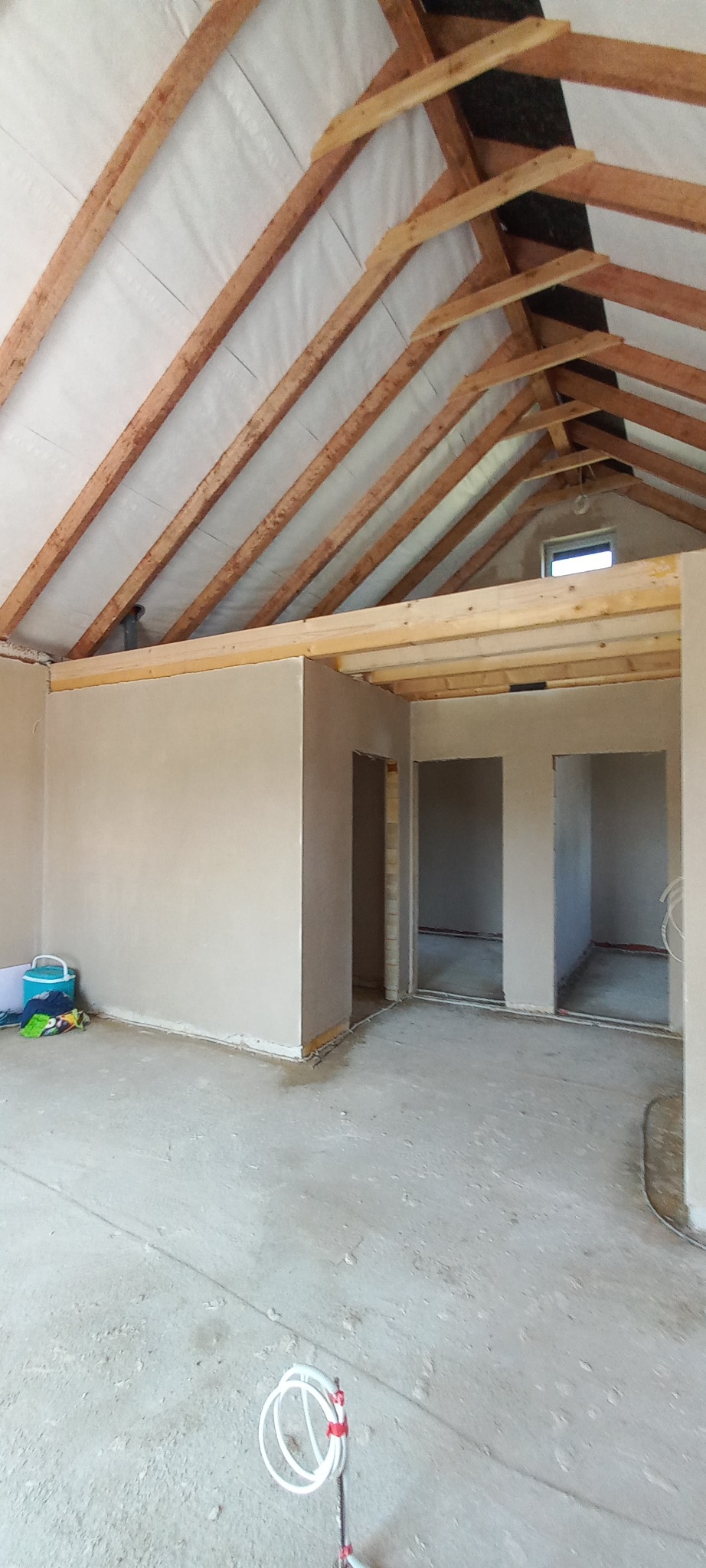 Surowe wnętrze poddasza z drewnianą konstrukcją dachu i świeżo otynkowanymi ścianami, przygotowane do dalszych prac wykończeniowych; widoczne otwory drzwiowe i narzędzia malarskie na podłodze.