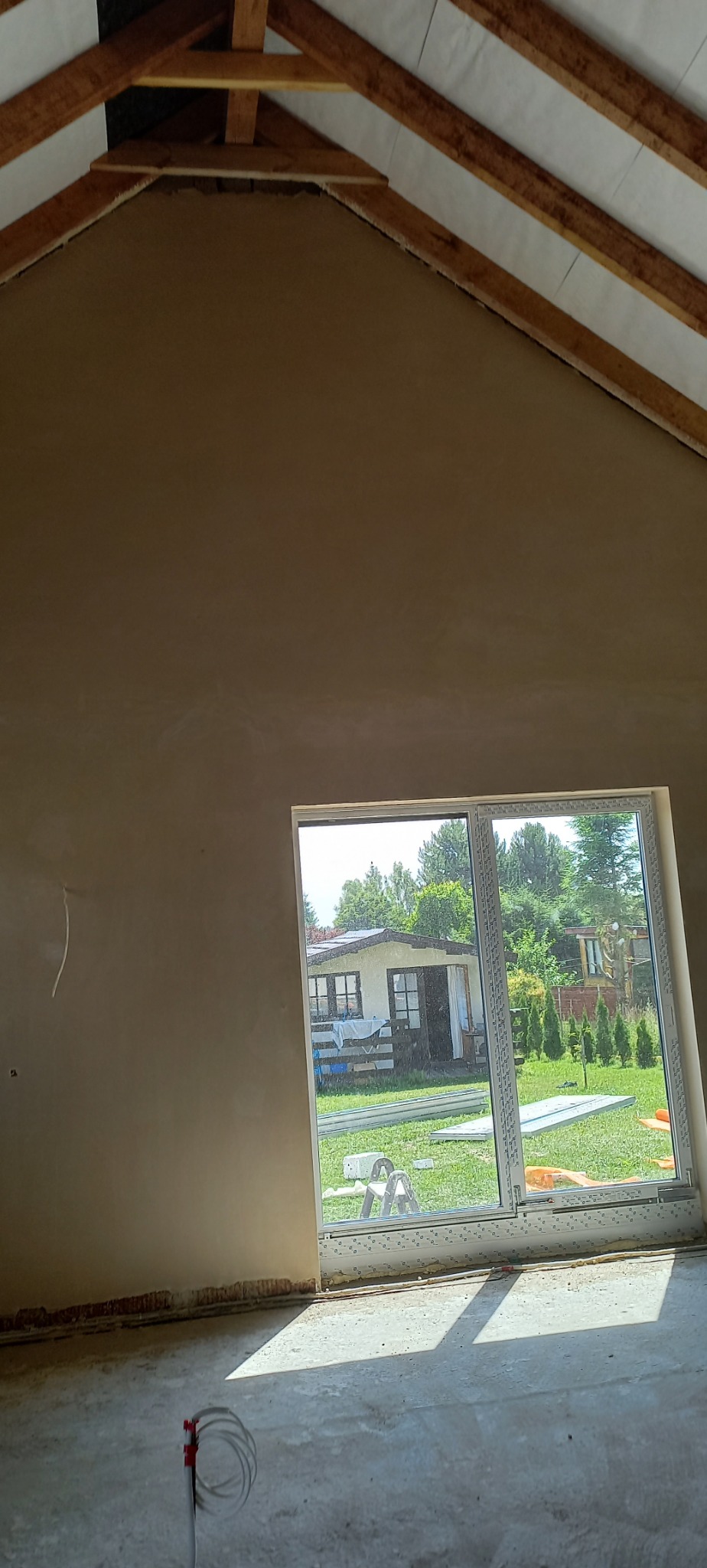Wnętrze poddasza z nałożonymi gładziami, widoczne krokwie dachowe, przez okno widok na ogród z domkiem.