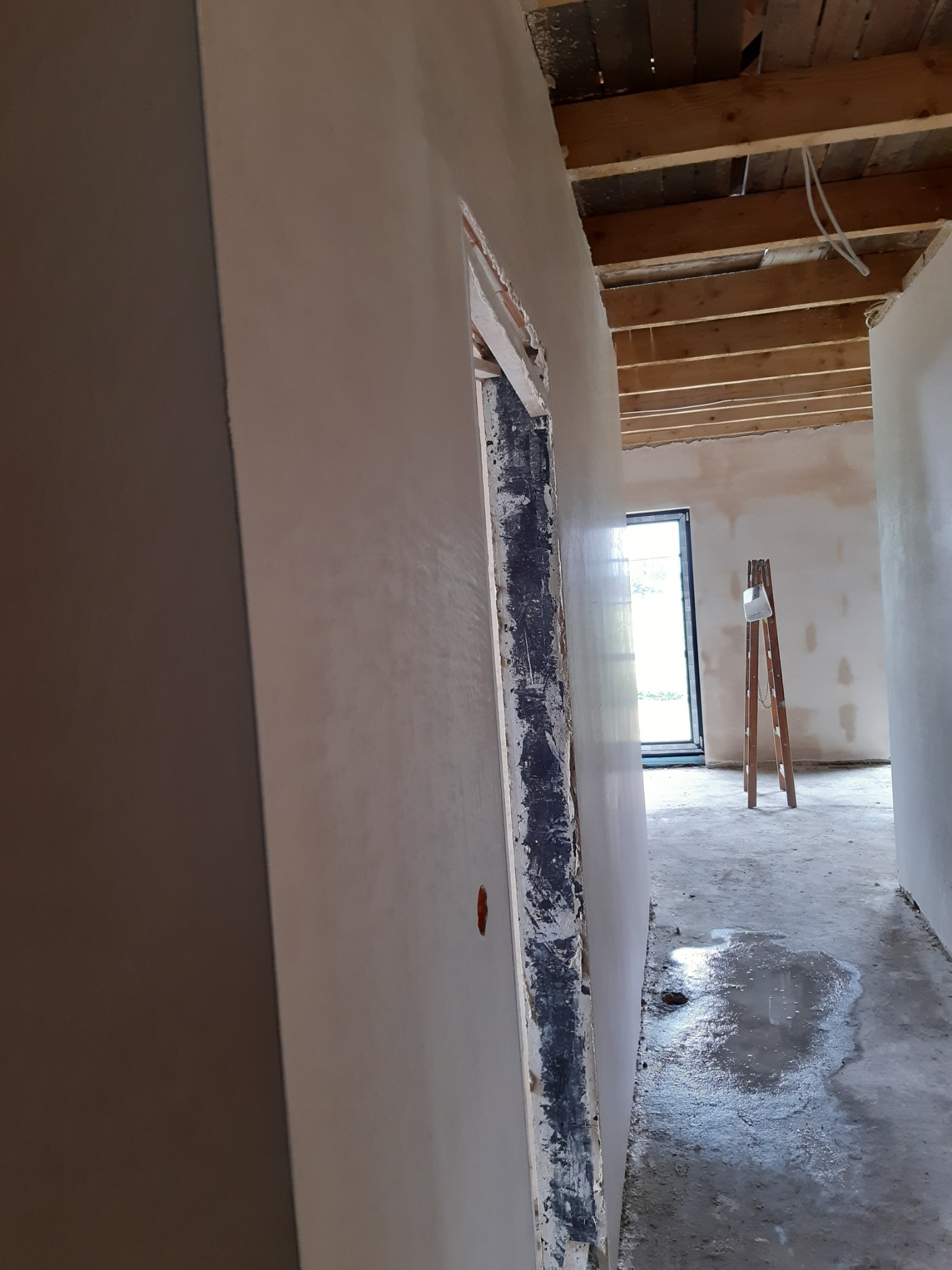 Świeżo otynkowane ściany w surowym wnętrzu, widoczne drewniane belki stropowe, na podłodze kałuża wody, drabina oparta o ścianę w tle.