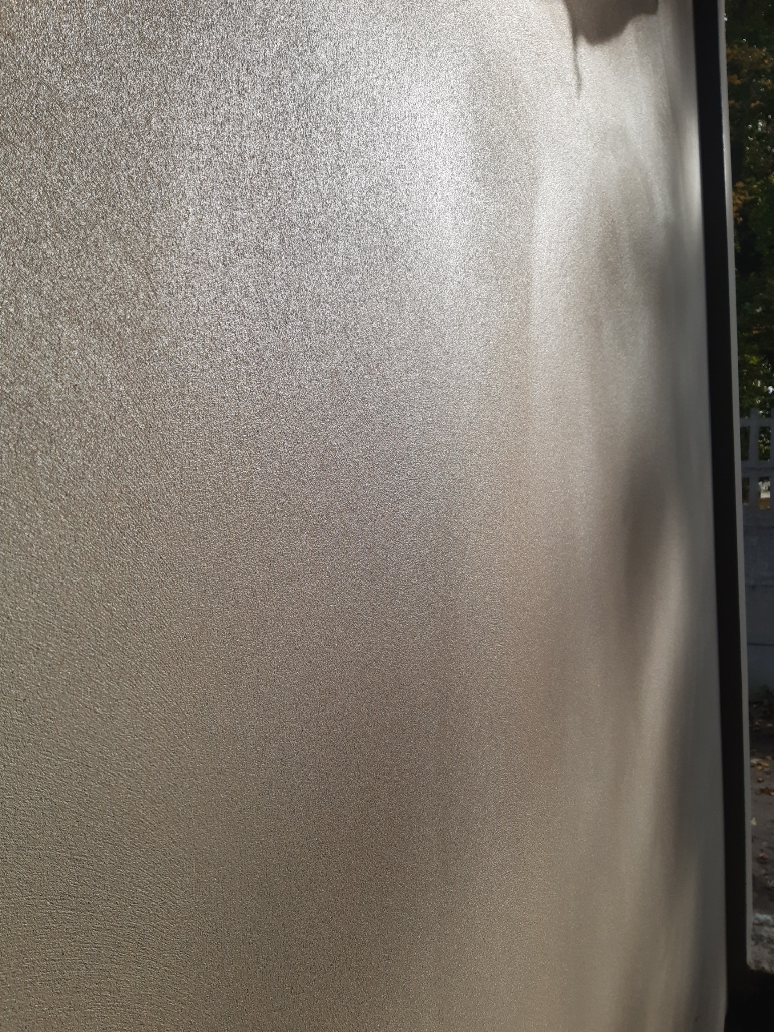 Świeżo nałożona, jasna, strukturalna powłoka na ścianie zewnętrznej budynku, z widocznym cieniem i fragmentem drzewa w tle.