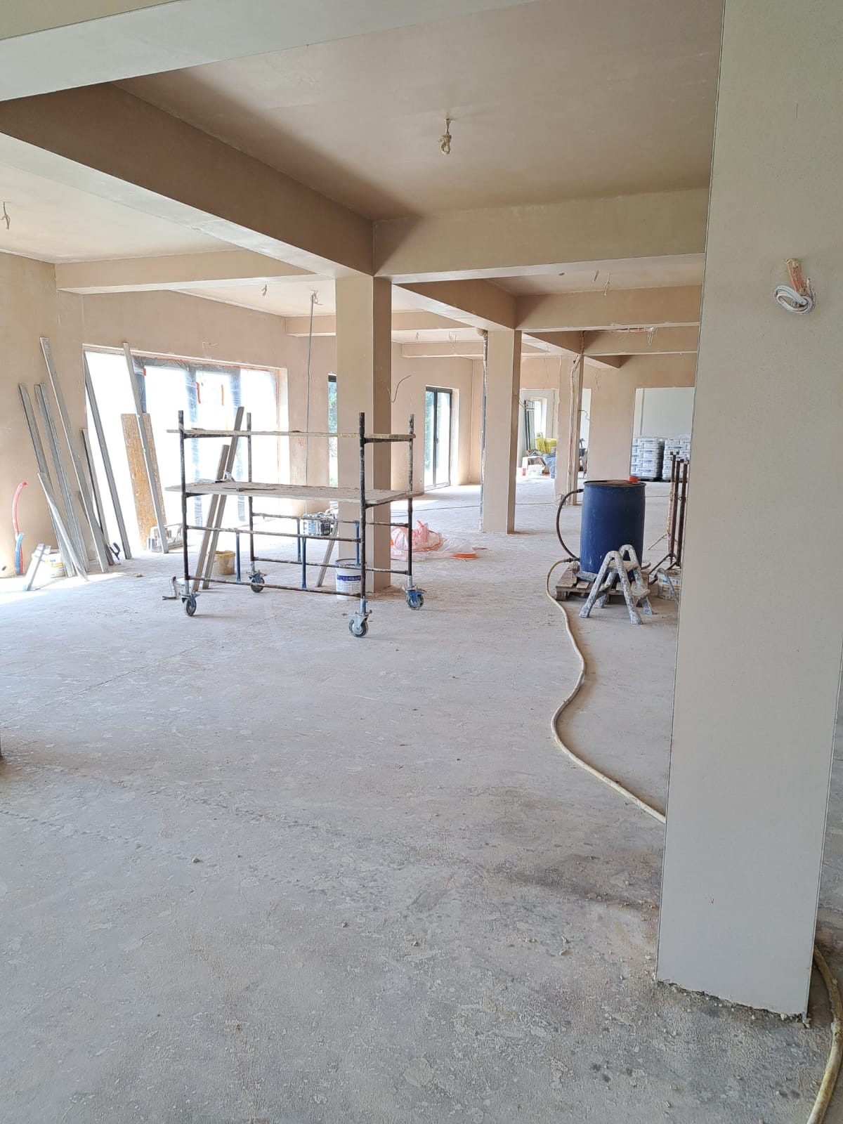 Wnętrze budynku w trakcie prac wykończeniowych; widoczne świeżo położone tynki na ścianach i suficie, metalowe rusztowanie na kółkach, błękitny pojemnik i przewód; surowy stan, przygotowanie...