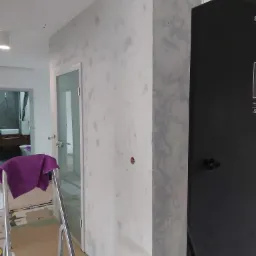 Ściana z nałożoną, ale jeszcze niewyszlifowaną gładzią w odcieniach szarości, widoczna drabina z fioletową szmatką, drzwi z szybą i fragment czarnej zabudowy meblowej.