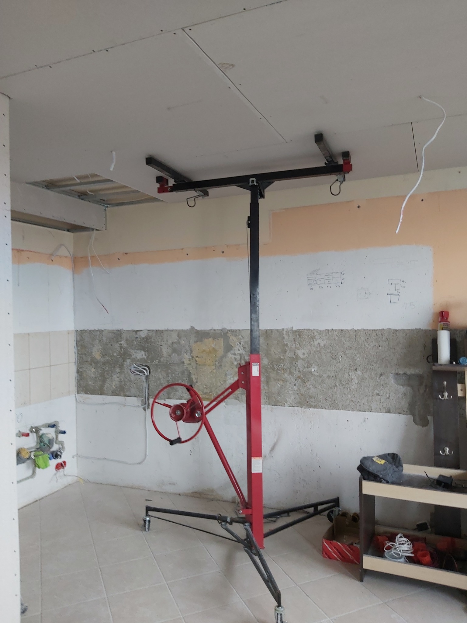 Wnętrze w trakcie remontu z częściowo zamontowaną płytą karton gips na suficie, widoczny podnośnik do płyt, odsłonięte instalacje na ścianach i szkice projektowe.