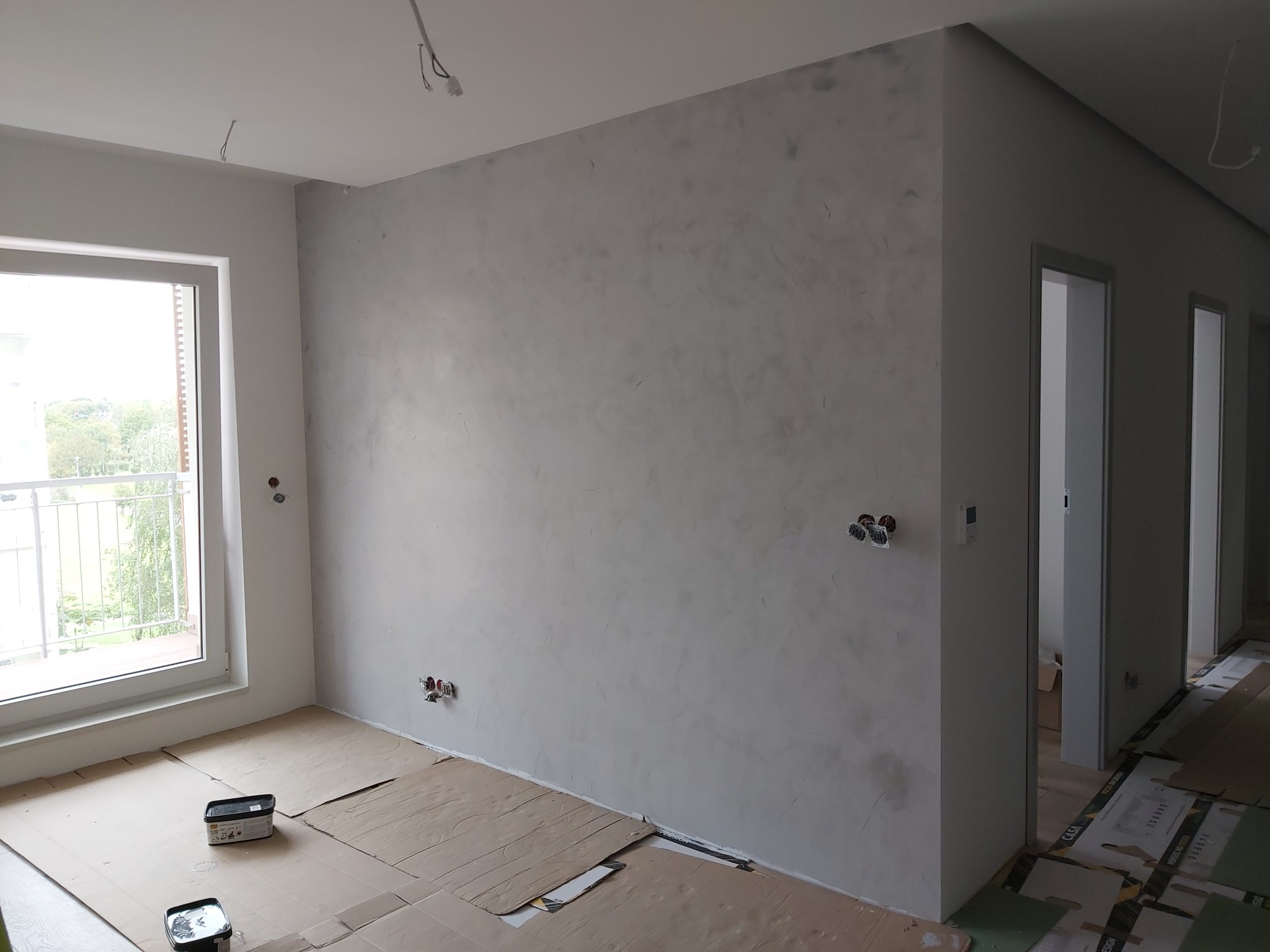 Ściana w pokoju po nałożeniu szarej, dekoracyjnej struktury, zabezpieczona podłoga kartonem, widoczne puszki elektryczne i narzędzia malarskie.