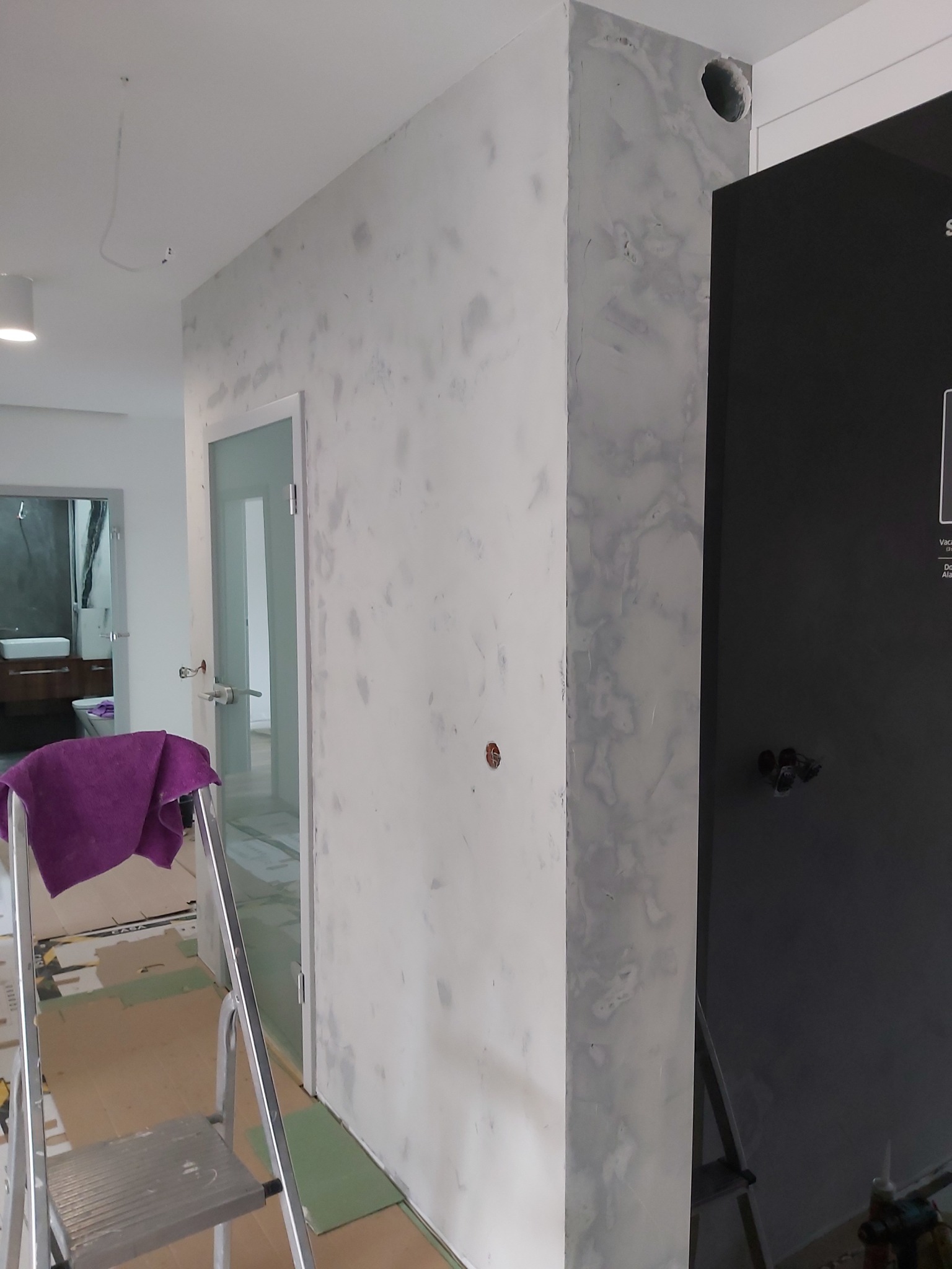 Ściana z nałożoną, ale jeszcze niewyszlifowaną gładzią w odcieniach szarości, widoczna drabina z fioletową szmatką, drzwi z szybą i fragment czarnej zabudowy meblowej.
