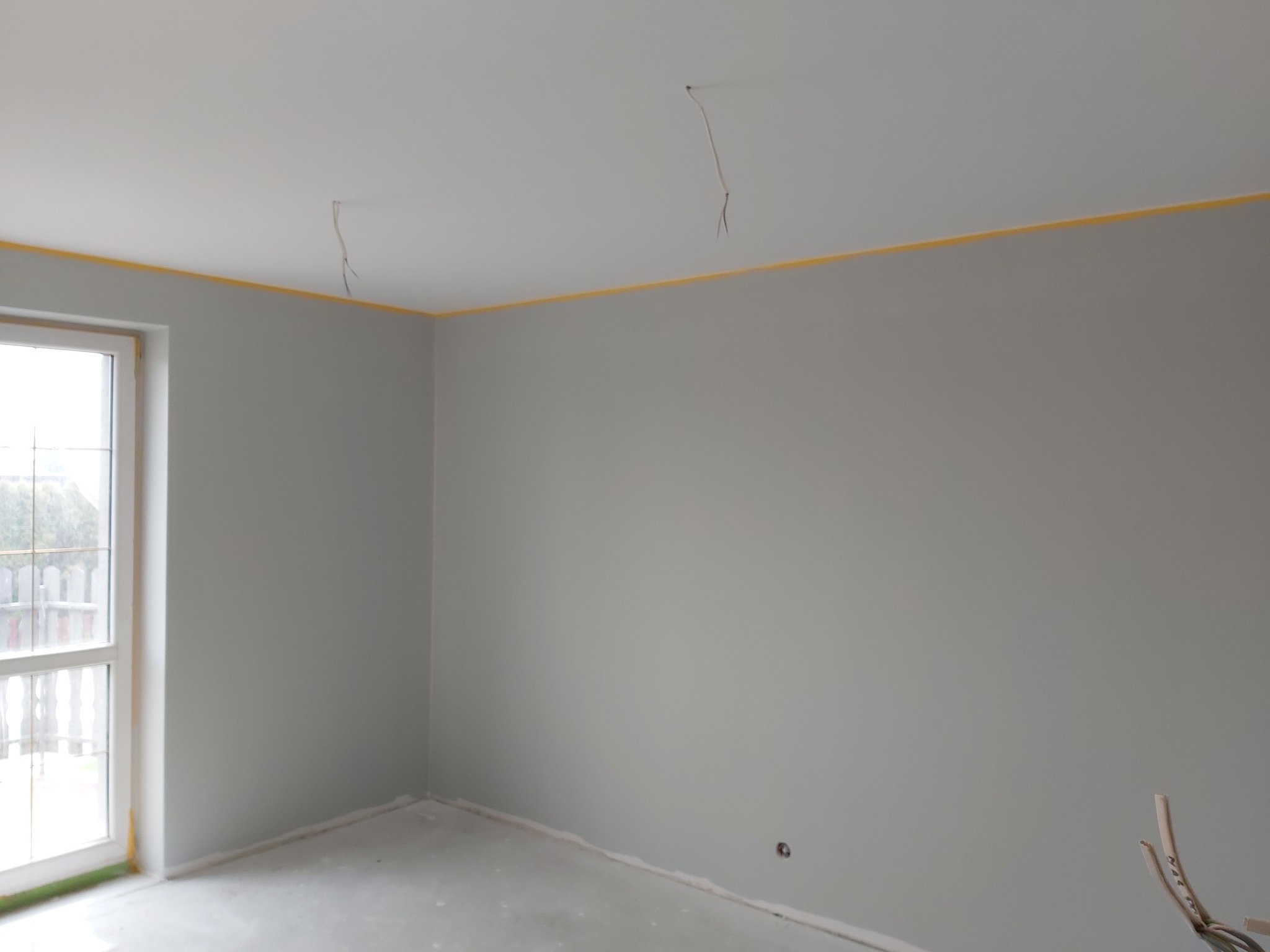 Pomalowane na szaro wnętrze pokoju w trakcie wykańczania, z widocznymi przewodami elektrycznymi wystającymi z sufitu i żółtą taśmą malarską zabezpieczającą krawędzie.