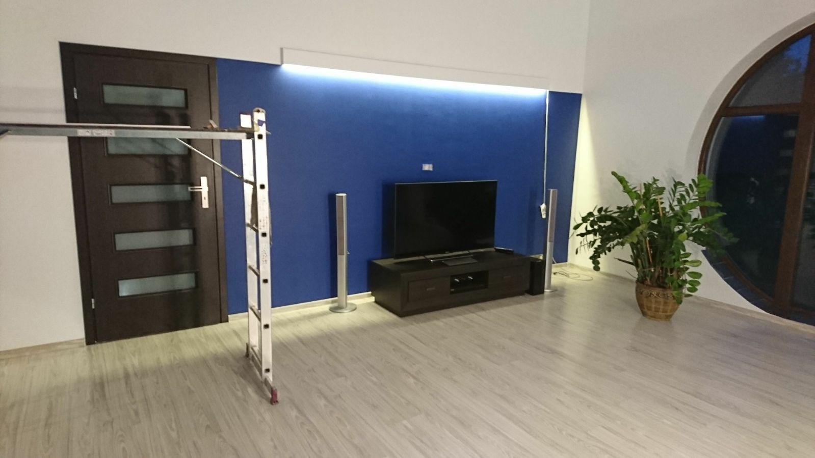 Pokój po częściowym odświeżeniu: pomalowana na niebiesko ściana, telewizor na szafce RTV, głośniki, roślina doniczkowa i drabina malarska przy brązowych drzwiach.