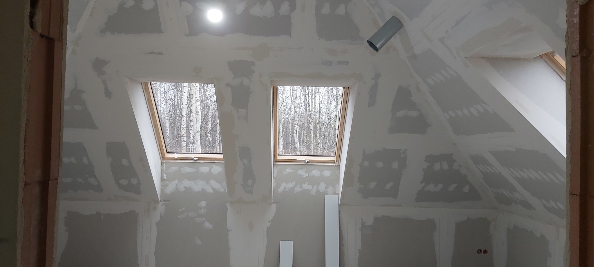 Poddasze w trakcie prac wykończeniowych, widoczne okna dachowe, ściany z płyt kartonowo-gipsowych ze śladami szpachlowania, sufit z widoczną instalacją wentylacyjną.