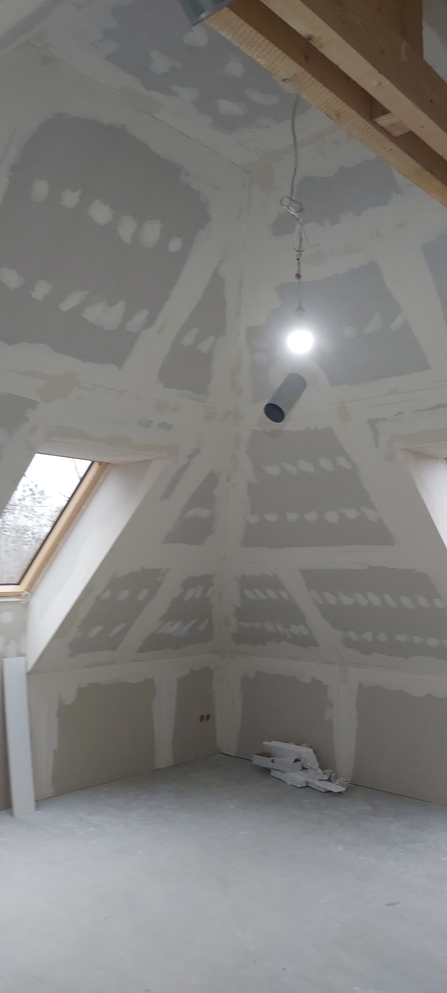 Poddasze ze skośnym sufitem w trakcie prac wykończeniowych, widoczne płyty kartonowo-gipsowe z widocznymi łączeniami wymagającymi szpachlowania i gładzi, surowe drewniane belki konstrukcyjne...