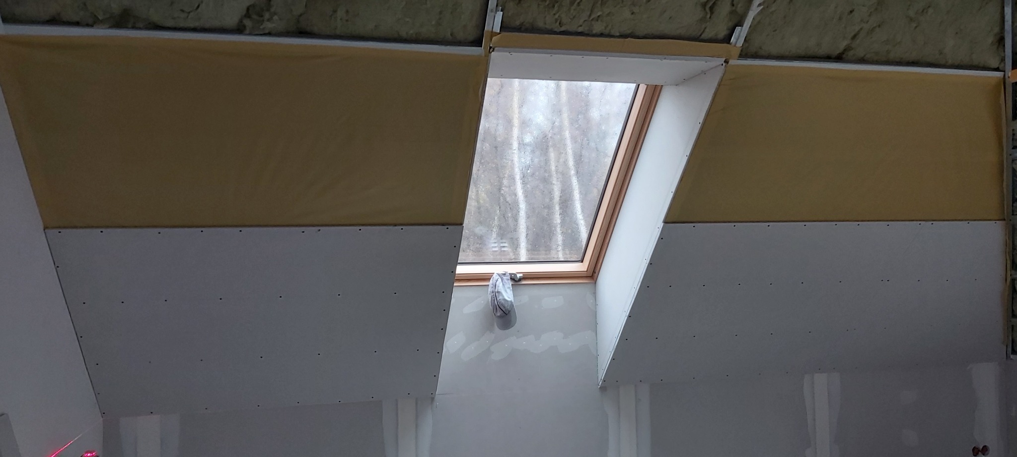 Montaż płyty karton gips na poddaszu z oknem dachowym, widoczne profile i izolacja, surowy stan przed szpachlowaniem, czapka wisząca na klamce okna.