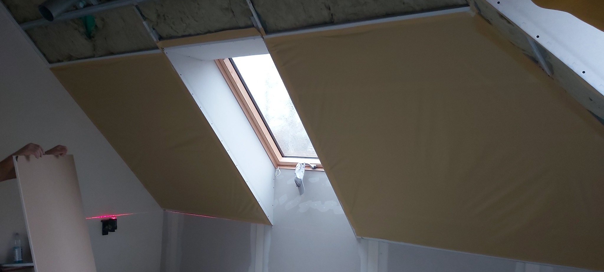 Montaż płyty karton gips na poddaszu z oknem dachowym, widoczny laserowy wskaźnik poziomu.