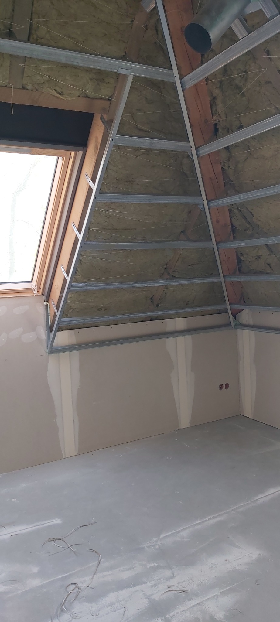 Strop poddasza z oknem dachowym w trakcie izolacji wełną mineralną i montażu metalowej konstrukcji pod płyty gipsowo-kartonowe, widoczne ślady gładzi na ścianach i podłodze.
